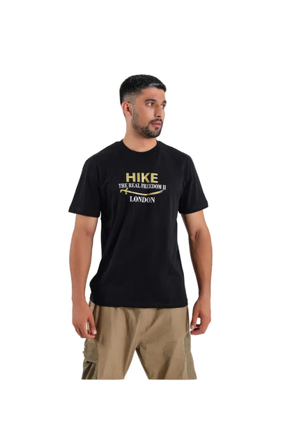 A man wearing a black short-sleeve t-shirt with a gold and white graphic print on the chest that reads 'HIKE THE REAL FREEDOM II LONDON' with a sword graphic, paired with khaki pants, against a light gray background.